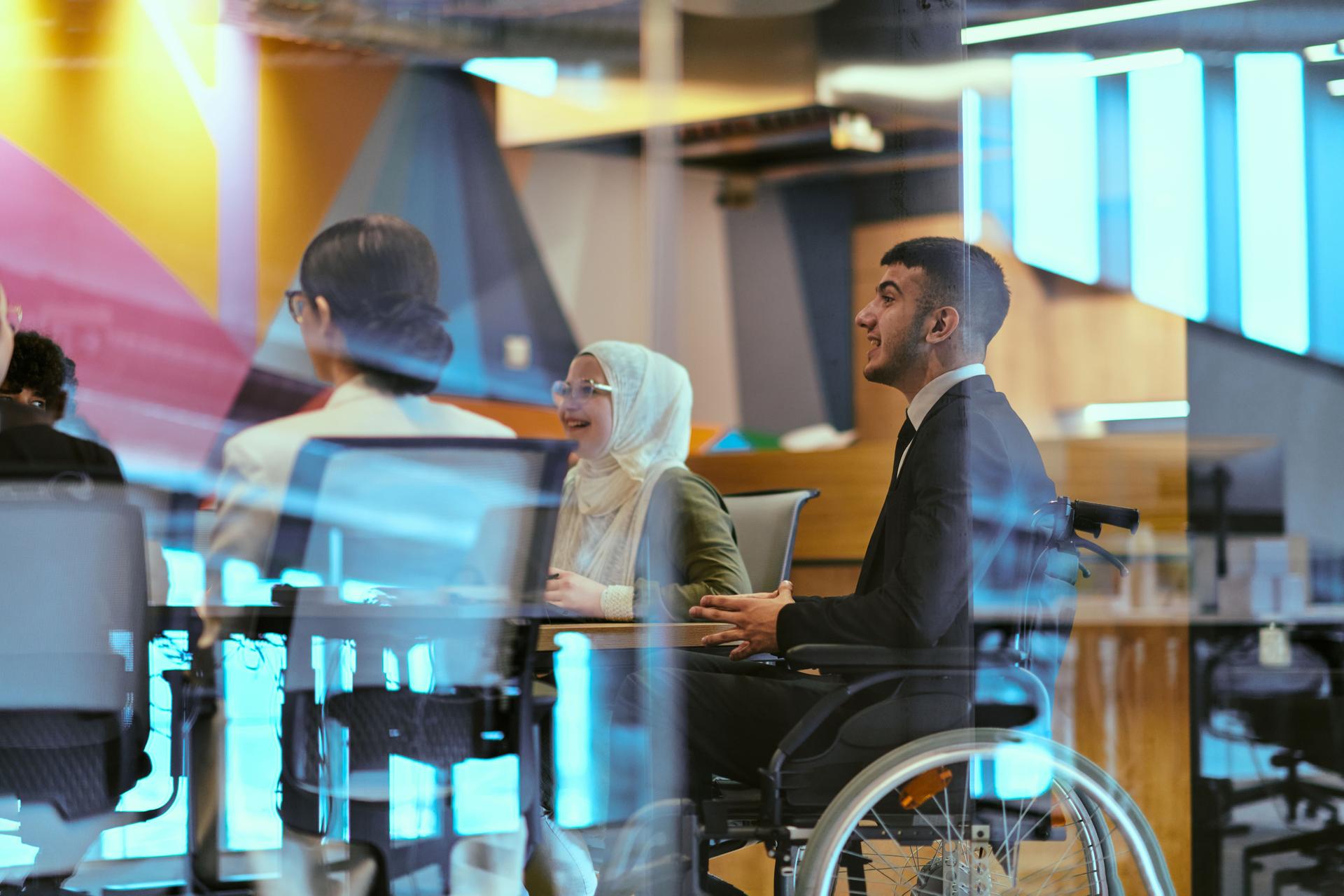 In a modern glass startup office, a wheelchair-bound director leads a successful meeting with colleagues, embodying inclusivity and teamwork.